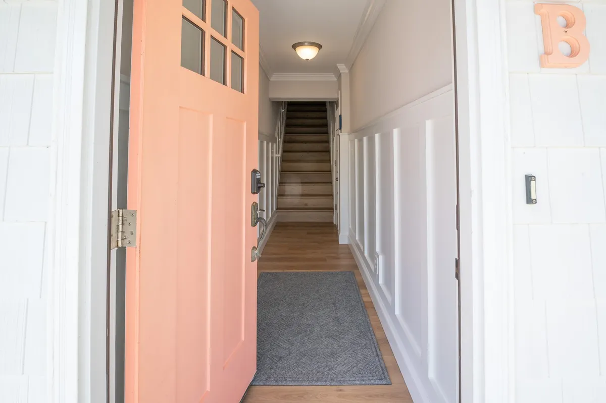 The photo features an orange front door leading into the home, a clean doormat, and a staircase beyond. The warm tones and neat entryway evoke feelings of arriving at a fresh, updated, and welcoming space.