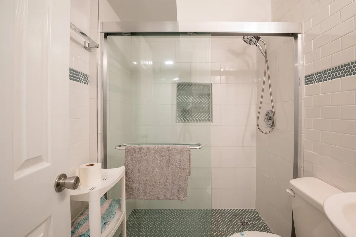 A clean, modern bathroom with a glass shower enclosure, herringbone tile floor, wall-mounted showerhead, towel rack with neatly arranged towels, toilet, and a shelf stocked with toiletries. The space feels fresh and inviting, ideal for daily routines.