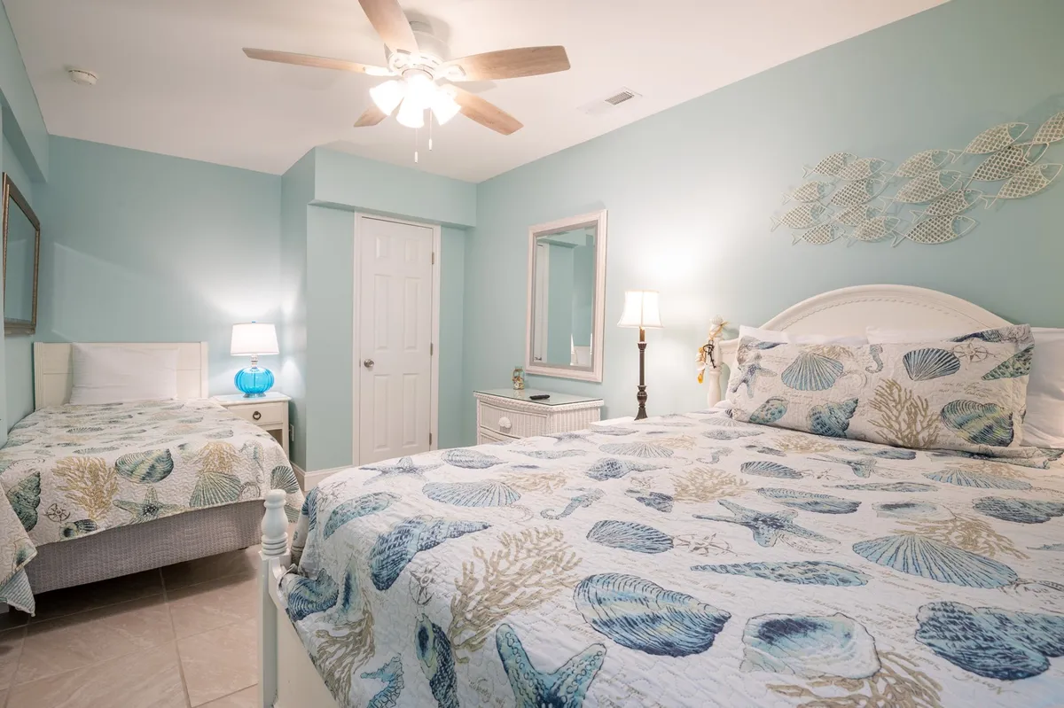 The photo showcases a room with both a large bed and a small twin bed, each dressed in seashell-patterned quilts. Additional elements include a blue bedside lamp, a mirror, and tasteful wall art, all bathed in natural sunlight.