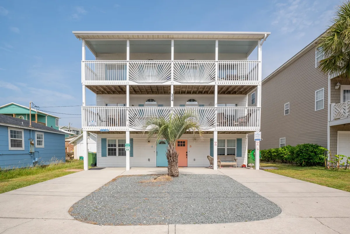 The image showcases the exterior of a cheerful Kure Beach condo, framed by a graceful palm tree, inviting balconies with white railings, and a pastel-colored front door. The scene radiates a welcoming, beachy vibe, with spaces perfect for relaxing outdoors and soaking in the coastal atmosphere.