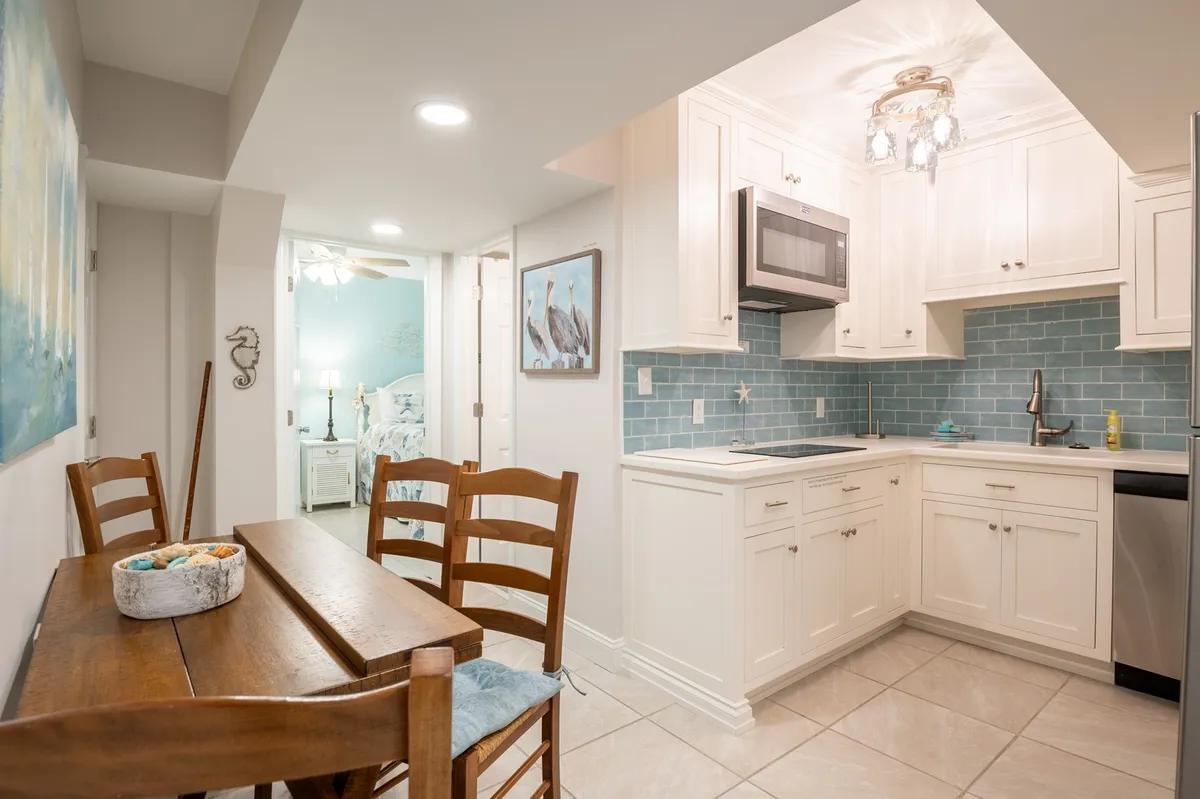 A cozy, open-concept living area featuring a farmhouse dining table set for four, modern kitchen cabinetry with a striking blue backsplash, and a prominent overhanging light fixture. The scene is set for both dining and relaxing, with ample natural light and thoughtful design touches that invite gathering and easy conversation.