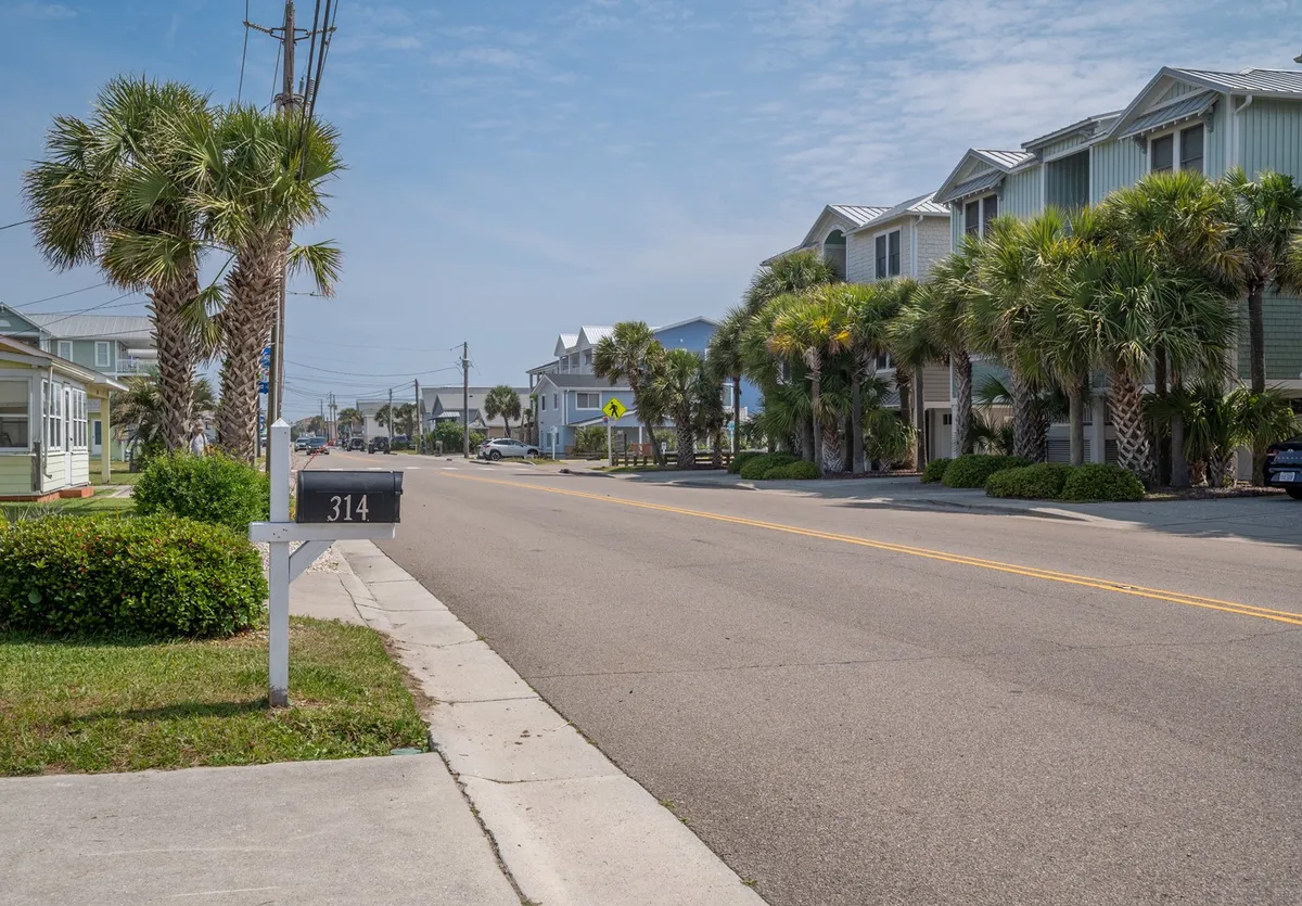 This exterior image captures a relaxed, palm-lined street with a welcoming mailbox numbered 314, rows of palm trees, and coastal houses. The scene evokes the feeling of a tranquil coastal neighborhood, inviting visitors to take leisurely strolls and explore the charming surroundings. The perspective hints at the proximity to the beach and local attractions, making it ideal for couples seeking romantic walks and access to dining or arts venues.