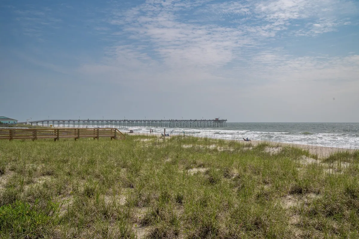 A tranquil scene featuring a long pier stretching into the ocean, with soft beach grass in the foreground. The beach appears spacious and uncrowded, while the pier offers opportunities for fishing, walks, and scenic ocean views, all bathed in calm, golden light.