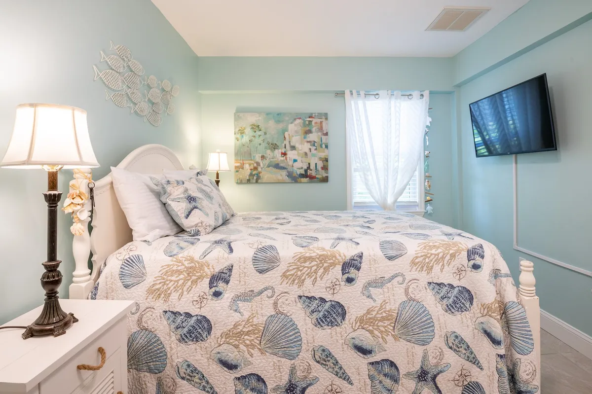 This image presents a tranquil bedroom scene featuring a comfortable bed adorned with a seashell-patterned quilt, a piece of wall art, a bedside lamp, and a wall-mounted TV. Gentle, natural light fills the room, creating a serene and restful atmosphere.