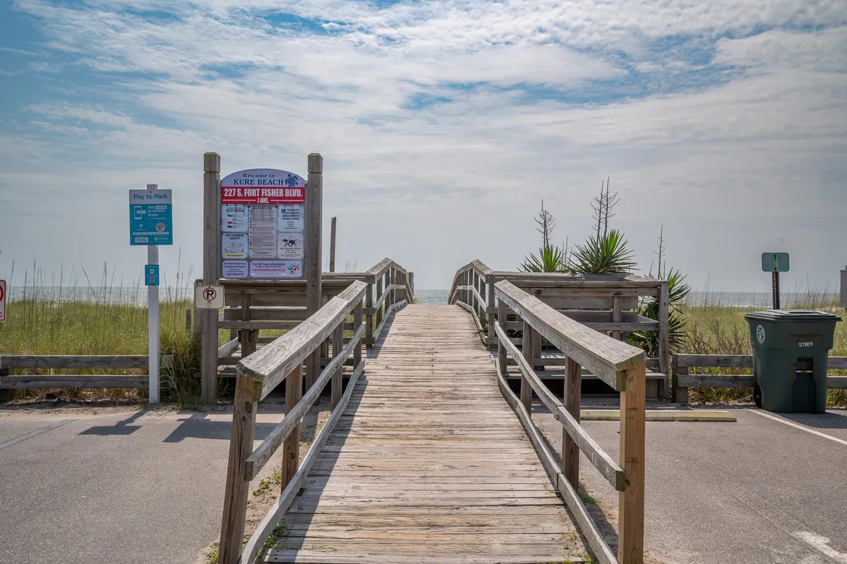 This image captures a wooden boardwalk leading directly toward the beach, framed by lush green grass and palm plants. Prominent is the beach access sign, signaling a welcoming entrance to the shoreline. The view suggests proximity and ease of access, with the boardwalk inviting visitors to take their first steps onto the sand.