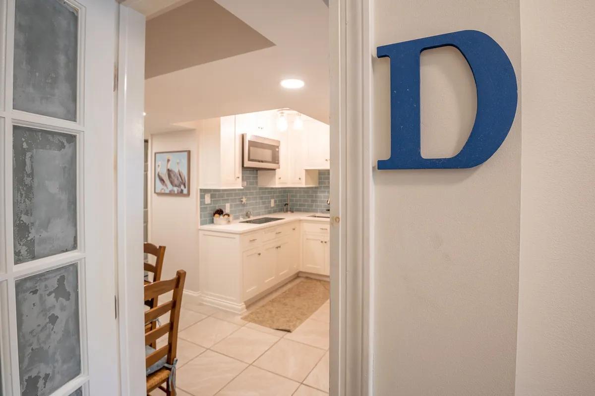 A welcoming kitchen and dining area with a full view of kitchen appliances, comfortable dining chairs, a distinctive blue letter 'D' as wall decor, and a soft blue tiled backsplash. The layout facilitates cooking, dining, and socializing all within one open space.