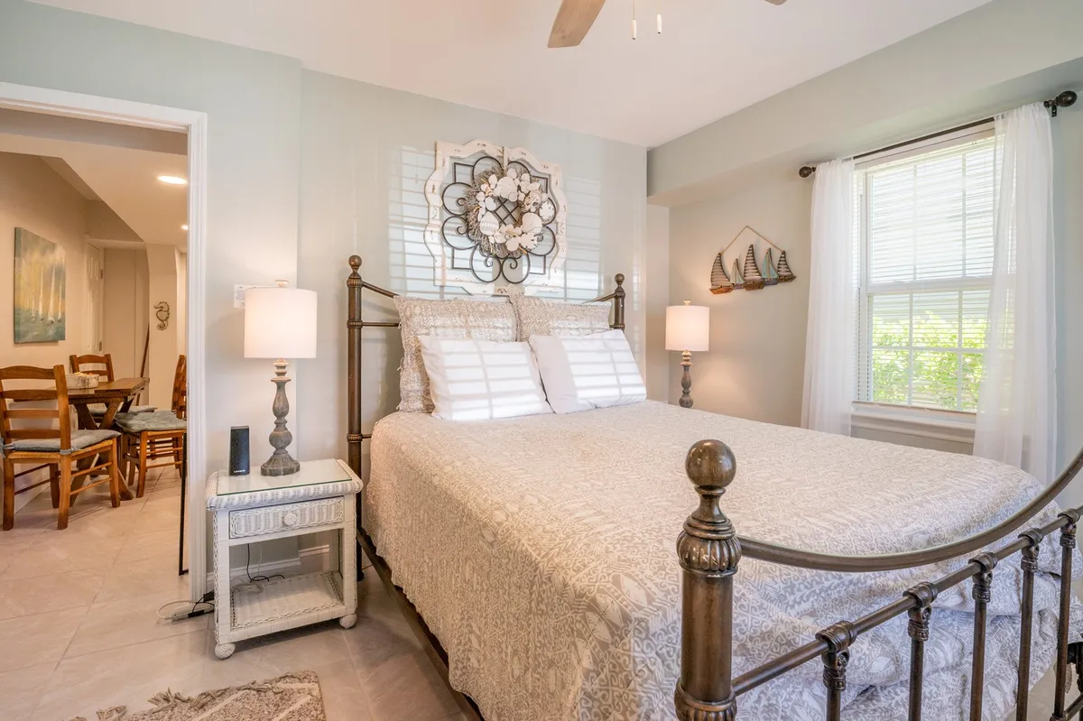 A beautifully appointed bedroom with a comfortable bed adorned with decorative bedding, flanked by nightstands and lamps. Sunlight streams through a window, casting a serene glow on the room and highlighting tasteful wall art above the bed.