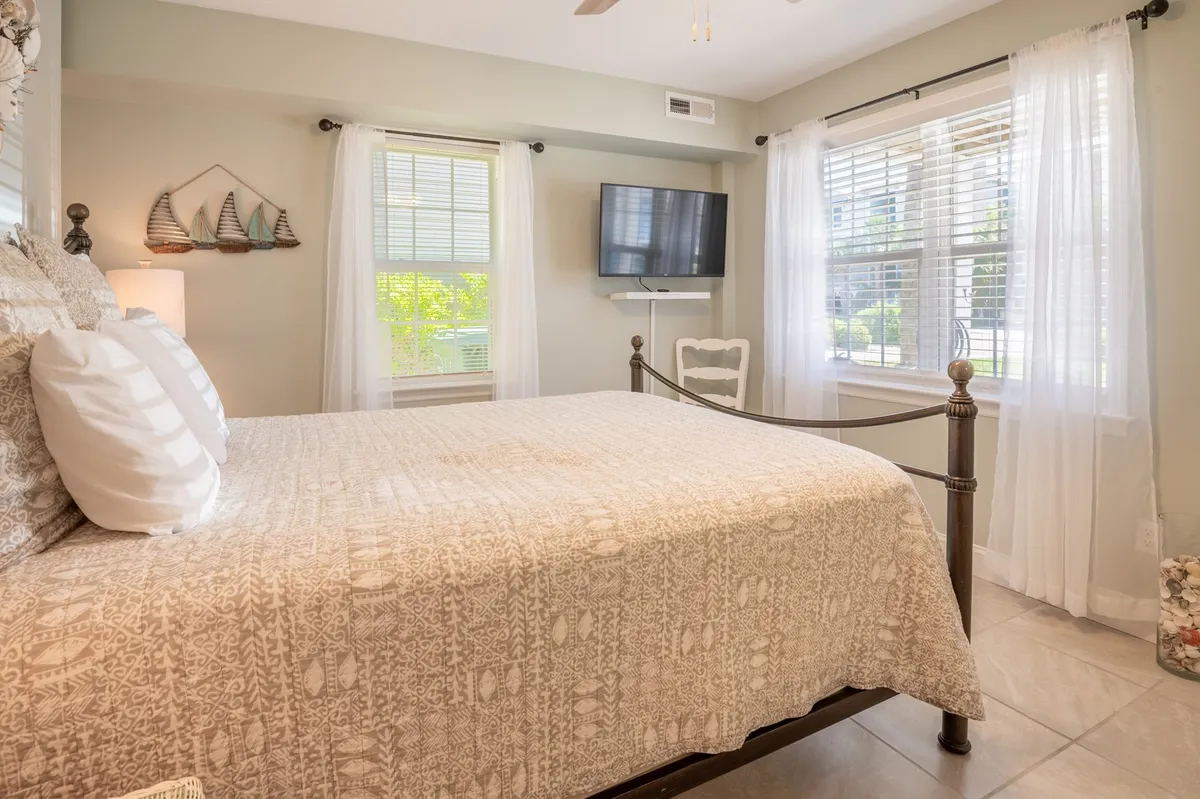 A peaceful bedroom scene featuring a neatly made bed, decorative boat artwork above the headboard, and large windows that allow natural light to fill the space. The ambiance is calm and inviting, evoking a sense of relaxation and comfort.